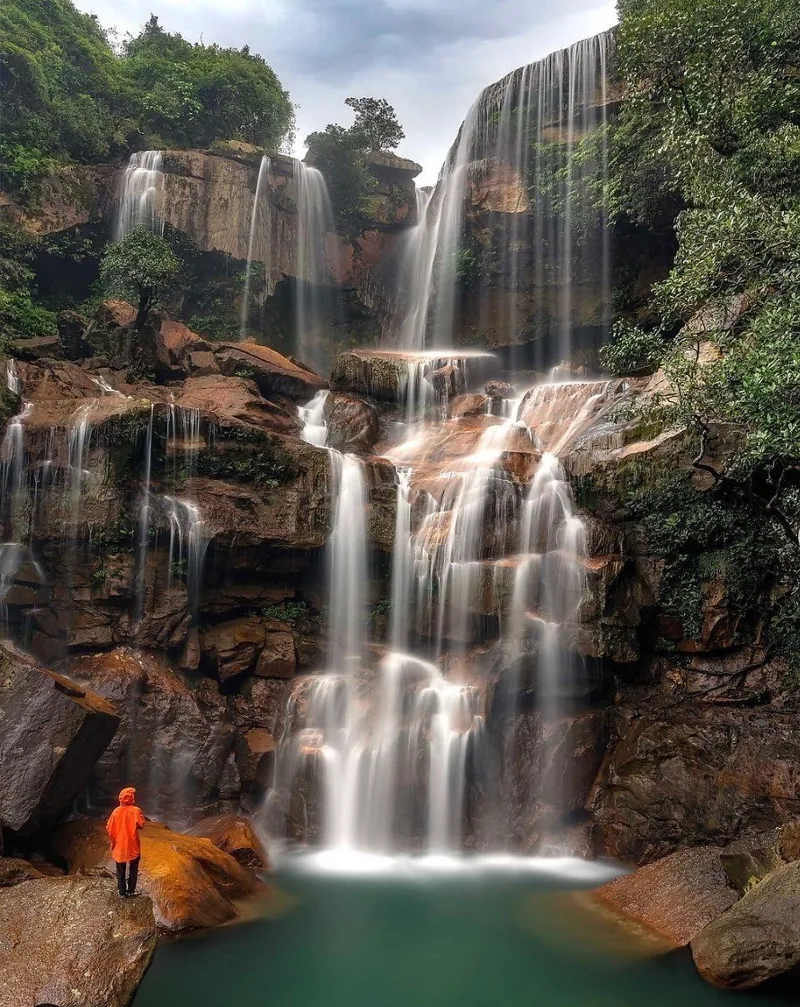 A trekker in an orange jacket stands before massive tiered waterfalls in the forests of Meghalaya