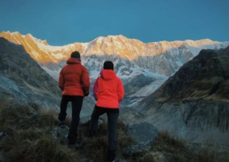 Two trekkers in orange and red down jackets standing on a ridge at sunrise, looking out at the snow-covered Annapurna massif