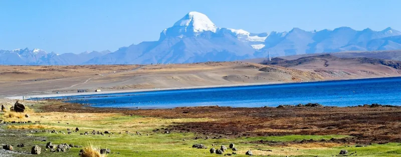 Mount Kailash with its snow-covered peak rising above Lake Mansarovar in the Tibetan plateau, blue sky and green foreground