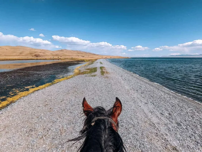 First-person view on horseback riding a narrow gravel strip between two channels of Son Kul Lake, Kyrgyzstan, with golden steppes and blue sky ahead