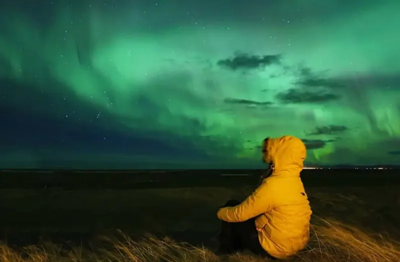 A traveller in a yellow jacket watching the northern lights blaze green across the Iceland sky