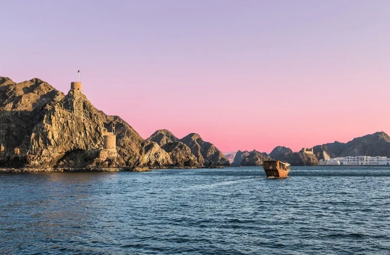 A traditional wooden dhow sailing past the Al-Jalali fort on Muscat harbour at sunset, rocky cliffs glowing pink behind it