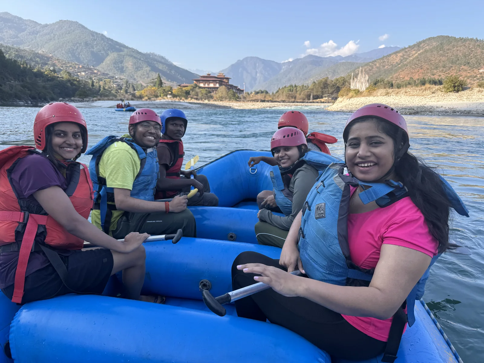 Trippers smiling in a blue raft on the river with Punakha Dzong behind
