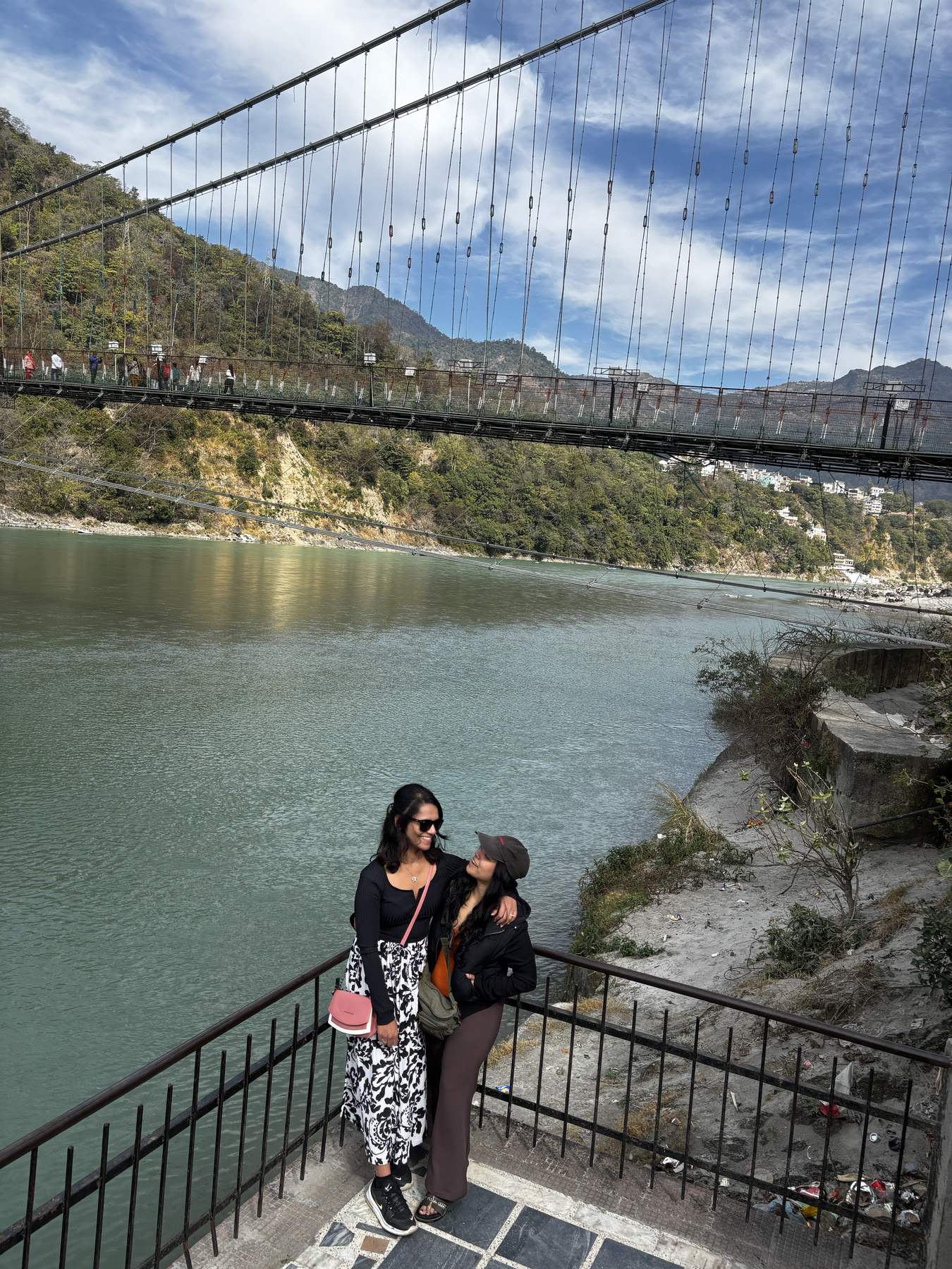 Two trekkers at a Rishikesh viewpoint with the Laxman Jhula suspension bridge over the Ganges behind