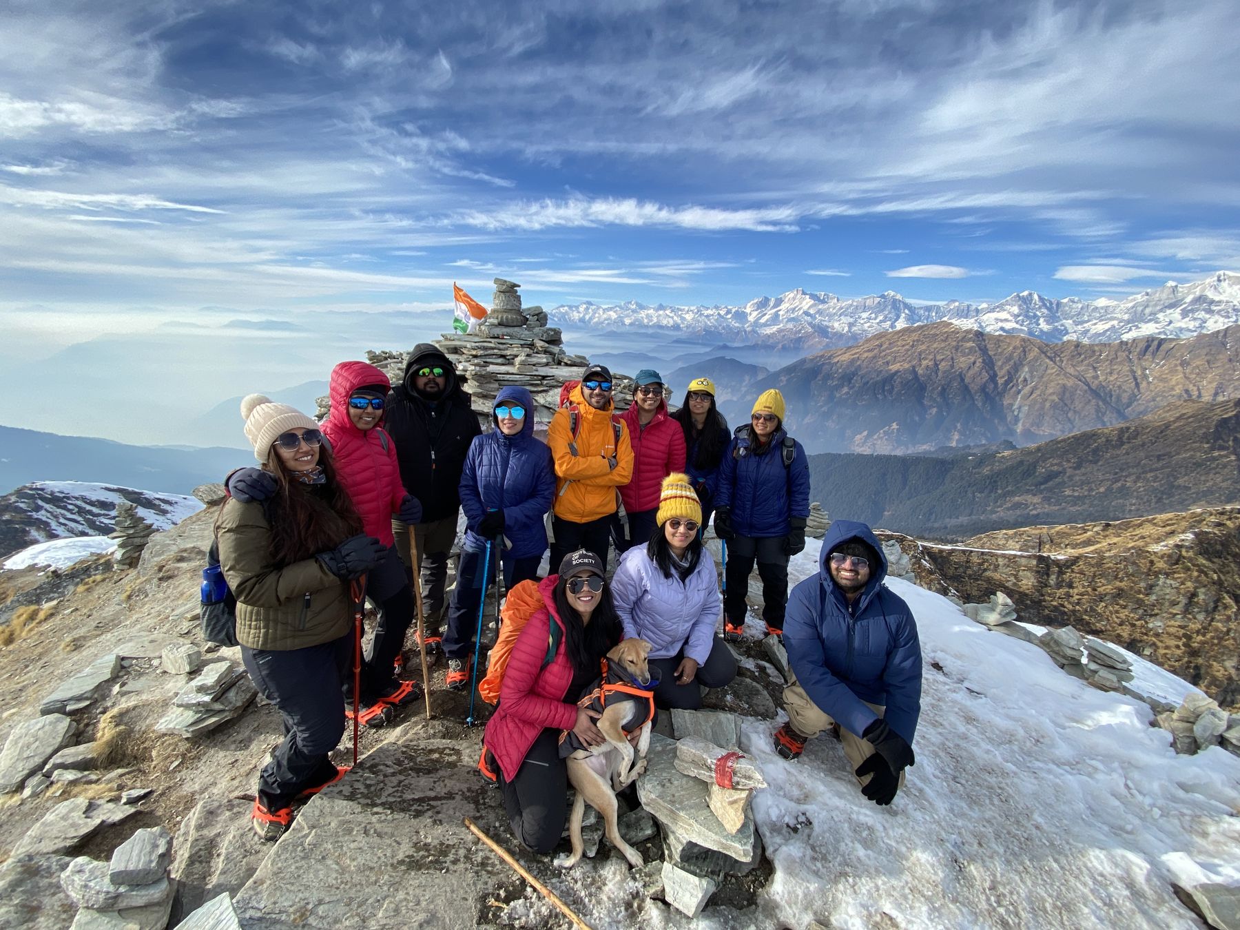 Trekkers on the snowy Chandrashila summit beside the stone cairn with Himalayan peaks behind