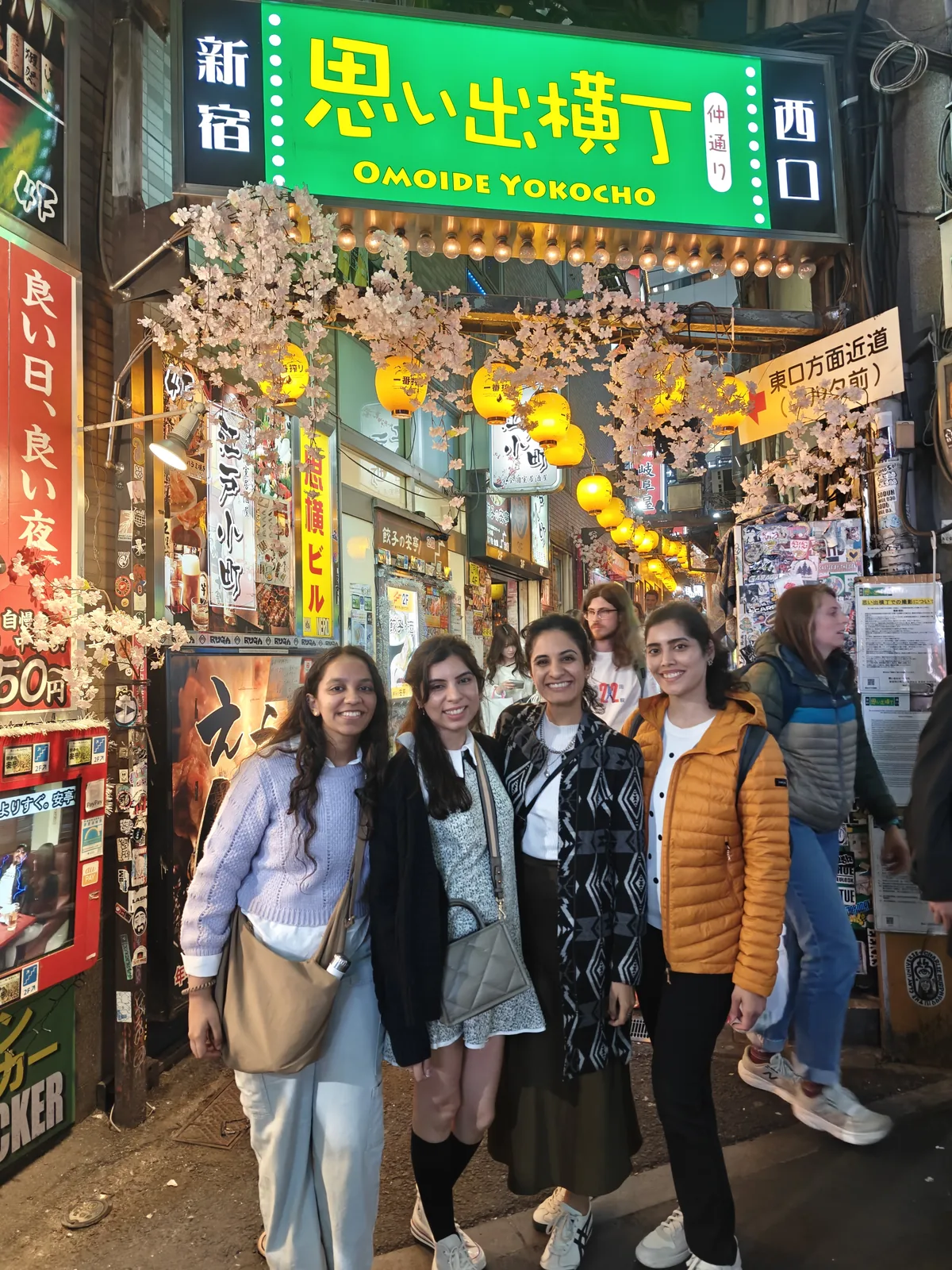 Japan Cherry Blossom, Day 2: Four trippers smiling at the Omoide Yokocho gate in Shinjuku, sakura decorations overhead