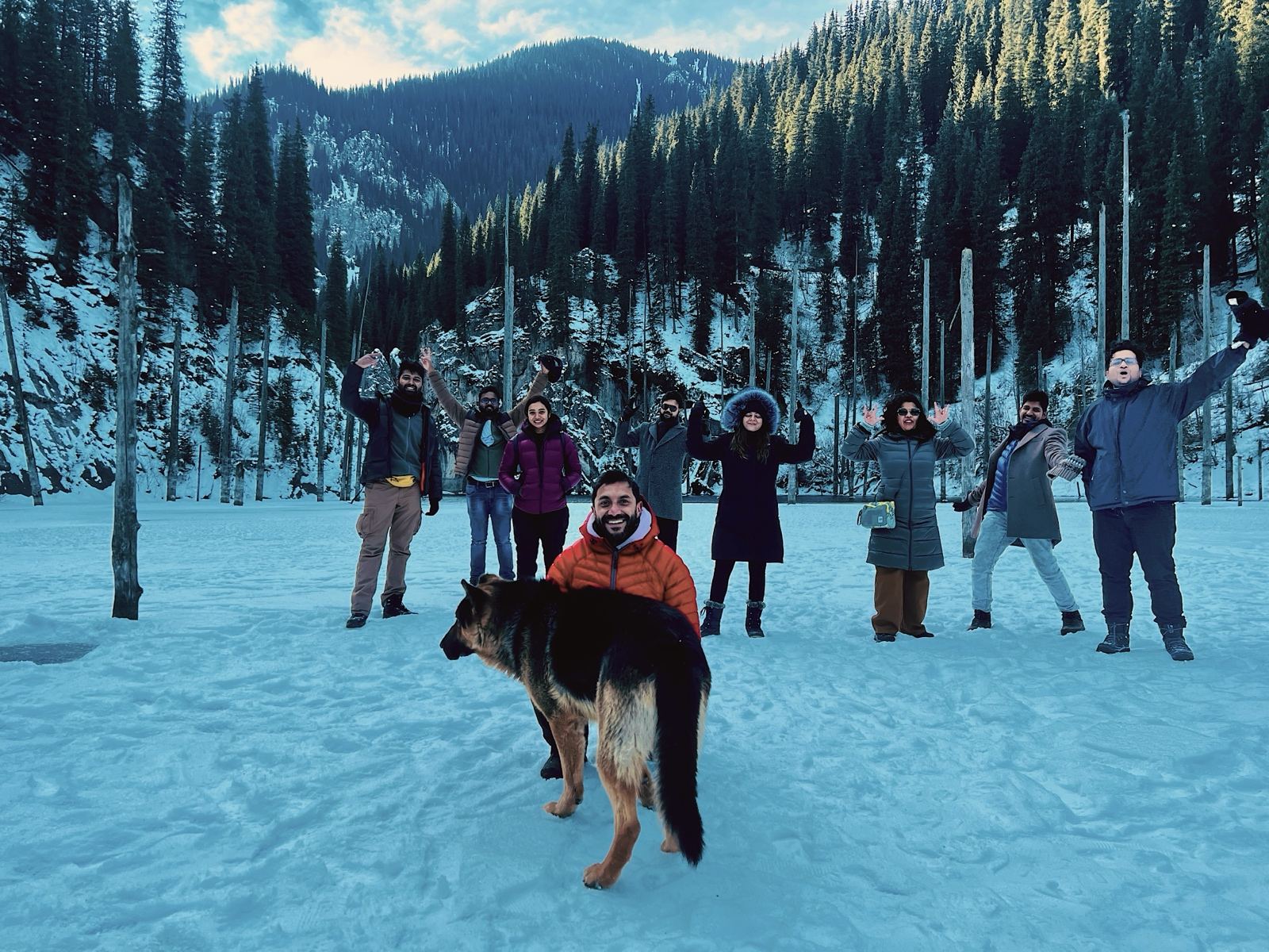 OJ group with arms raised on frozen Kaindy Lake, spruce trunks behind and a dog in front