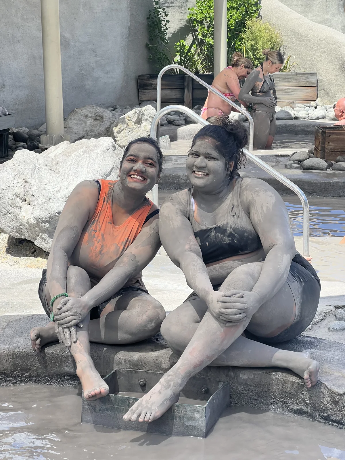 Two OJ trippers covered in grey mud, laughing on the edge of a Rotorua mud pool