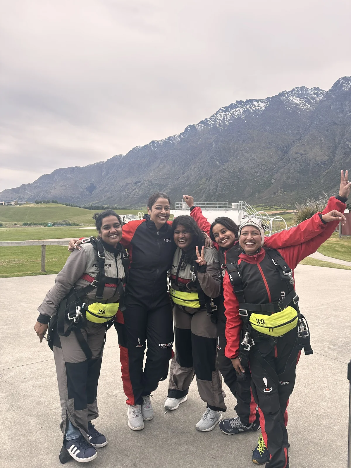 Five OJ trippers in skydiving jumpsuits and harnesses celebrating after the jump in Queenstown